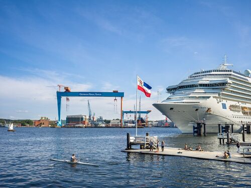Kreuzfahrtschiff am Anleger in Kiel mit Blick auf die Werft German Naval Yards und Menschen am Steg bei sonnigem Wetter