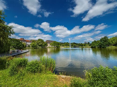 See mit Springbrunnen in einem Park, umgeben von Bäumen und Gebäuden unter blauem Himmel mit Wolken