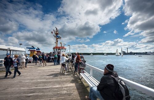 Menschen auf einem Holzsteg neben einem Hafen mit mehreren Segelbooten und einem großen Schiff unter bewölktem Himmel