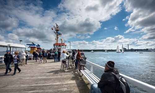 Menschen auf einem Holzsteg neben einem Hafen mit mehreren Segelbooten und einem großen Schiff unter bewölktem Himmel